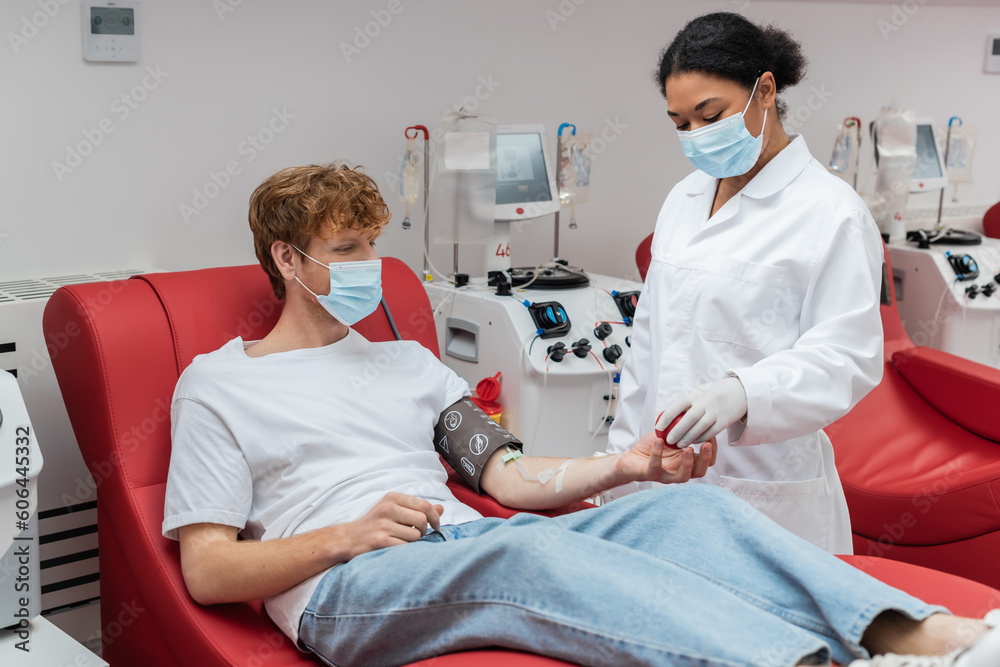 Obraz premium multiracial doctor in medical mask giving rubber ball to redhead donor in blood pressure cuff sitting on medical chair near transfusion machine in blood donation center