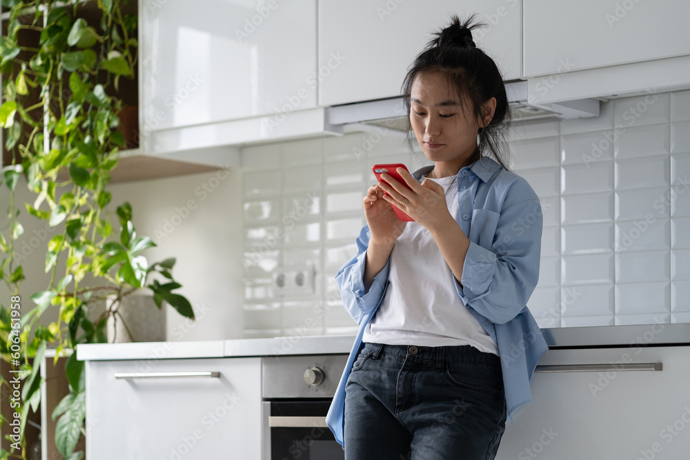 Ταπετσαρία Optimistic Asian woman stands in kitchen looks social media posts of friends and family with slight smile