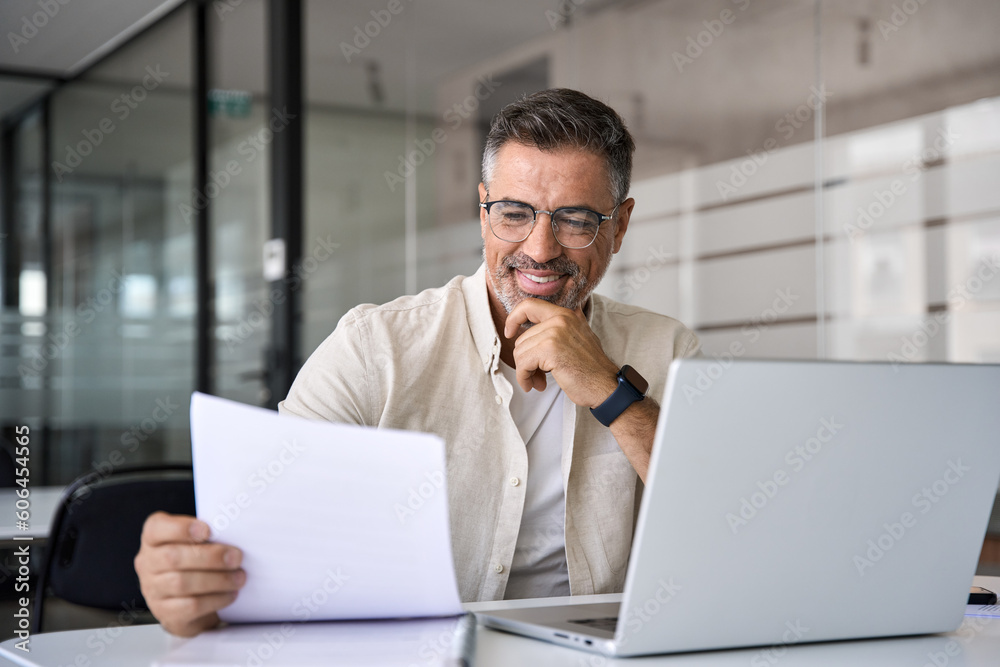 Smiling Latin or Indian male business man accountant analyst holding documents, work at laptop computer doing online trade market tech research. Focused Hispanic businessman with paperwork in office. Smiling Latin or Indian male business man accountant analyst holding documents, work at laptop computer doing online trade market tech research. Focused Hispanic businessman with paperwork in office.