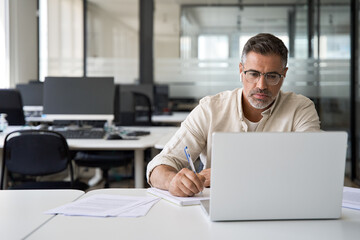 Middle-age Hispanic man using laptop computer for business studying, watch online virtual webinar training meeting, video call. Focused mature Indian or Latin businessman work in office, copy space.