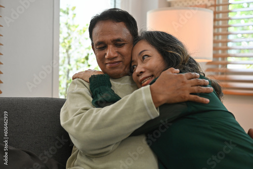 A retired Asian couple sweetly looks into each other's eyes as they embrace each other lovingly on the sofa in their home.