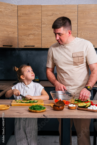 Cute little girl and her handsome young dad are cutting vegetables and smiling while cooking in kitchen at home.