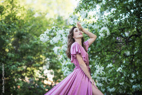 A young beautiful brunette girl stands near a branchy spring apple tree. A girl in a pink dress smiles between blooming apple trees. The concept of spring or summer