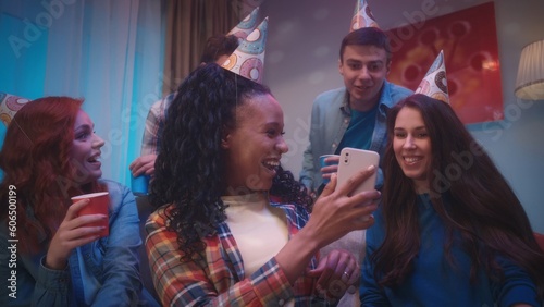 An African American woman shows the phone to her friends, they look at photos, videos. A group of friends in party hats in a room decorated for a party, with disco ball reflections on the walls.