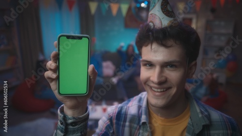 Curly guy in a festive hat, smiling, shows a phone with a green screen. Close up of a guy with a smartphone on a blurred background of a party, in a room decorated with a disco ball, flags.