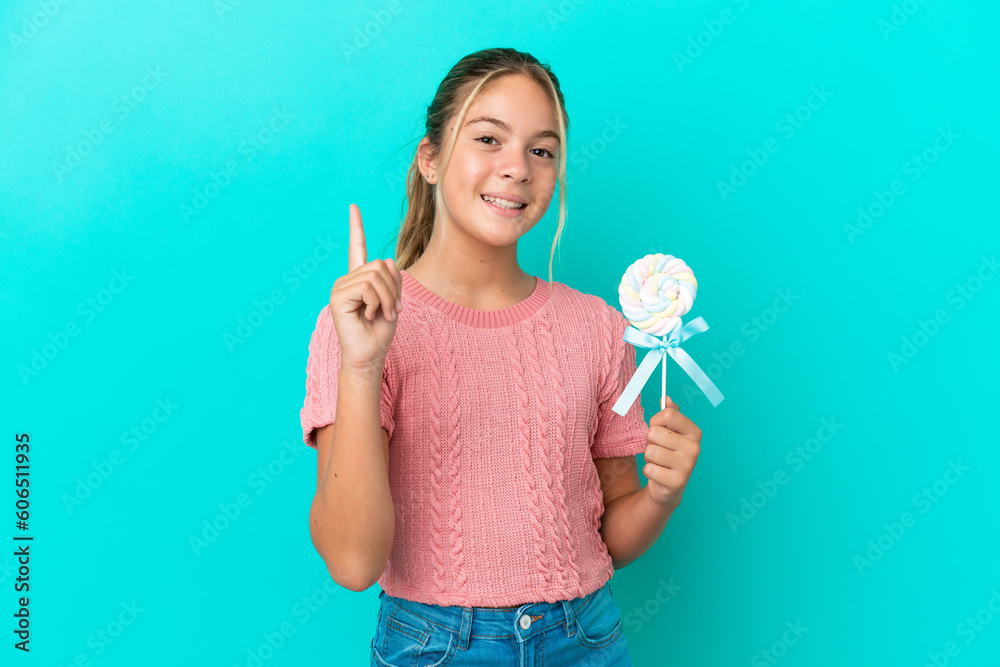 Little Caucasian girl holding a lollipop isolated on blue background showing and lifting a finger in sign of the best Little Caucasian girl holding a lollipop isolated on blue background showing and lifting a finger in sign of the best