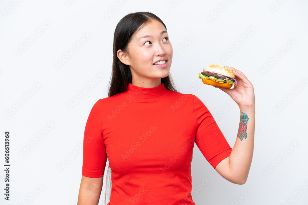 Young Asian woman holding a burger isolated on white background looking up while smiling Young Asian woman holding a burger isolated on white background looking up while smiling