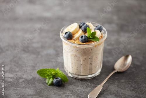 Healthy diet breakfast. Overnight oatmeal with chia seeds, bananas, peanut butter and blueberry in a glass jar on a gray concrete background.