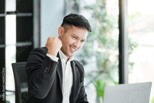 Successful and happy young Asian man, businessman, happy, shows with hand yes. Made a contract. Won money. Sitting with a smart phone or notebook at the table in a modern office.