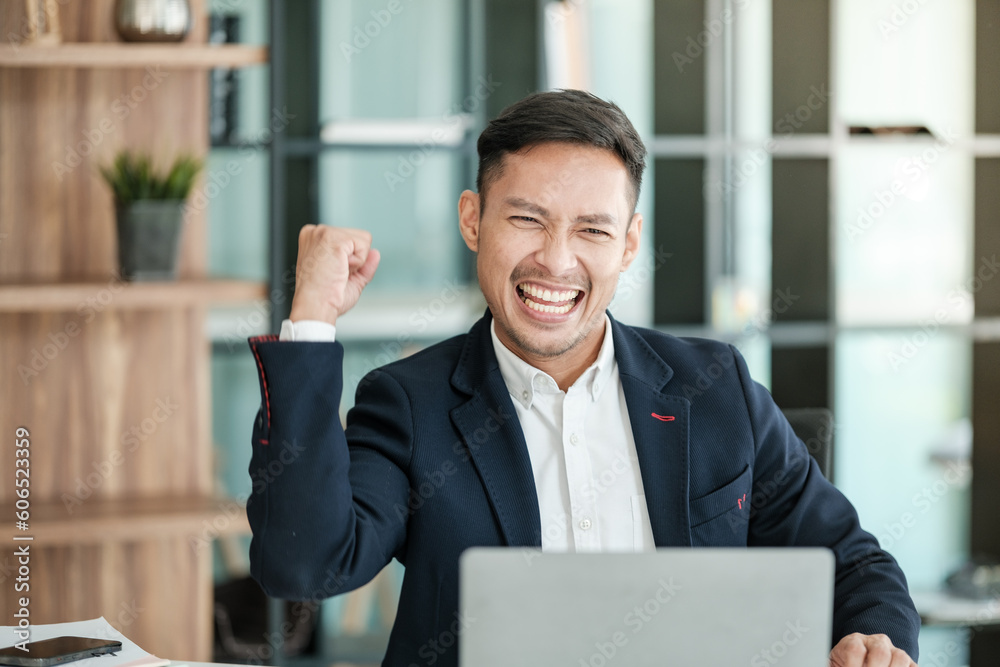 Successful and happy young Asian man, businessman, happy, shows with hand yes. Made a contract. Won money. Sitting with a smart phone or notebook at the table in a modern office. Successful and happy young Asian man, businessman, happy, shows with hand yes. Made a contract. Won money. Sitting with a smart phone or notebook at the table in a modern office.