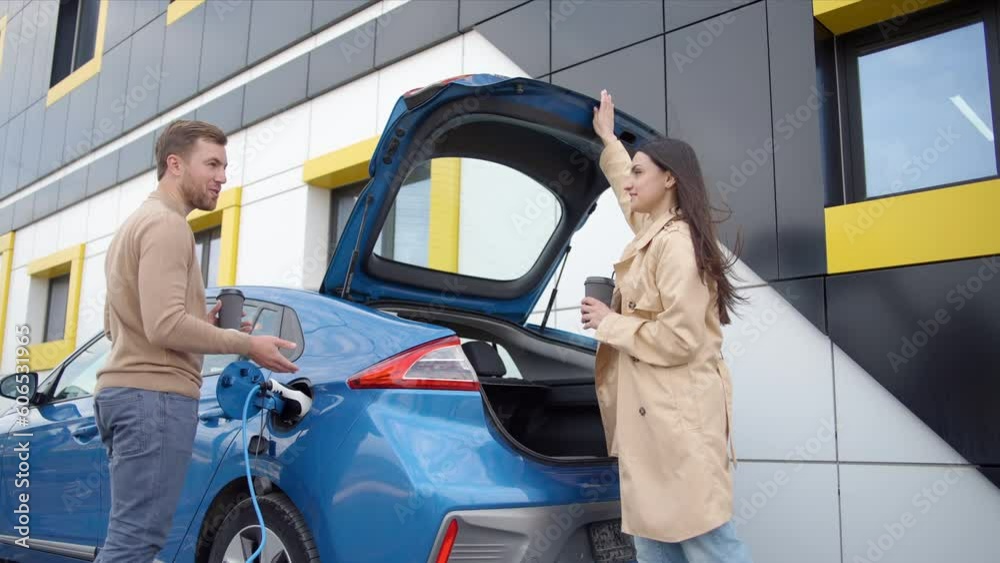 Couple of man and woman install charger plug from charging station to electric car before driving around city center. Girl with coffee near the trank of car. Eco friendly rechargeable car powered by c Couple of man and woman install charger plug from charging station to electric car before driving around city center. Girl with coffee near the trank of car. Eco friendly rechargeable car powered by c
