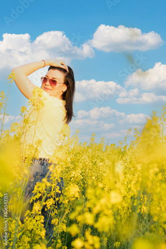 A beautiful girl in sunglasses stands in a field of yellow flowers against a blue sky, smiles happily and inhales the fresh spring and clean air. Peace concept. The color of the flag of Ukraine. © Fedash
