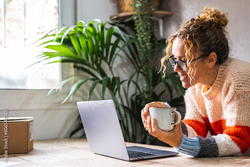 Happy adult woman using laptop at home and drinking coffee. Modern lady smiling at the computer screen in video chat connection leisure online activity. People at home surfing the net and enjoying day photo