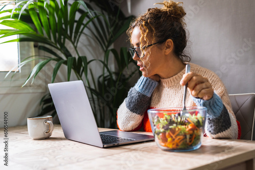 People eating fast and healthy in front of a computer during job lunch break. Modern business lifestyle working at home. One woman eat salads from transparent bowl and use notebook pc on the table