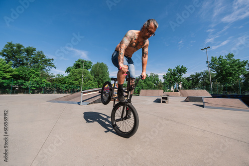A mature tattooed bmx rider is balancing on one wheel in a skate park.