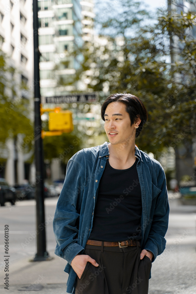 Portrait of attractive smiling asian man holding hands in pockets wearing stylish casual outfit looking away standing on the street