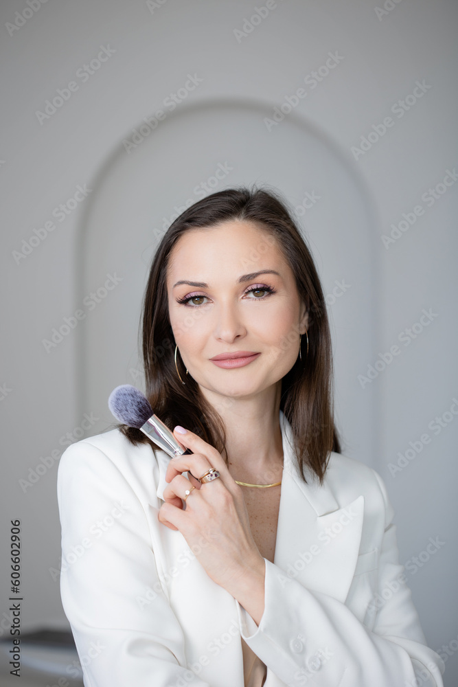 Fashion and beauty, stylish makeup artist in a white jacket with makeup brushes in her hands, business portrait of a makeup artist stylist or director of a beauty studio, arches on the background Fashion and beauty, stylish makeup artist in a white jacket with makeup brushes in her hands, business portrait of a makeup artist stylist or director of a beauty studio, arches on the background