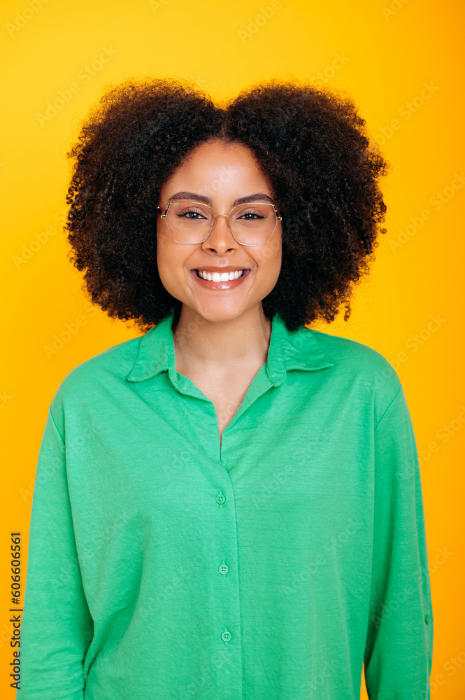 Vertical photo of a positive beautiful hispanic or brazilian curly haired woman, with glasses, wearing a green casual shirt, looking at the camera, smiling, standing on an isolated yellow background Vertical photo of a positive beautiful hispanic or brazilian curly haired woman, with glasses, wearing a green casual shirt, looking at the camera, smiling, standing on an isolated yellow background