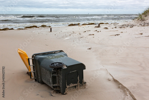 A green and yellow trash bin on a sandy ocean beach dotted with clumps of dune grass under a cloudy sky after a storm