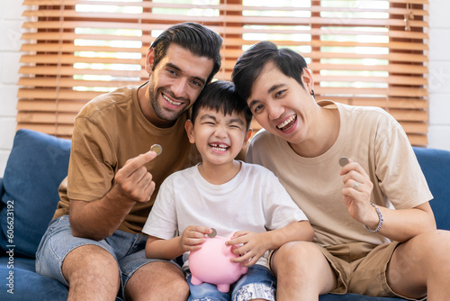 Two fathers and smiling son putting coin into piggy bank. Smiling boy sitting on father lap saving money in piggybank. Gay couple teaching son to save money while putting coin in piggy bank.