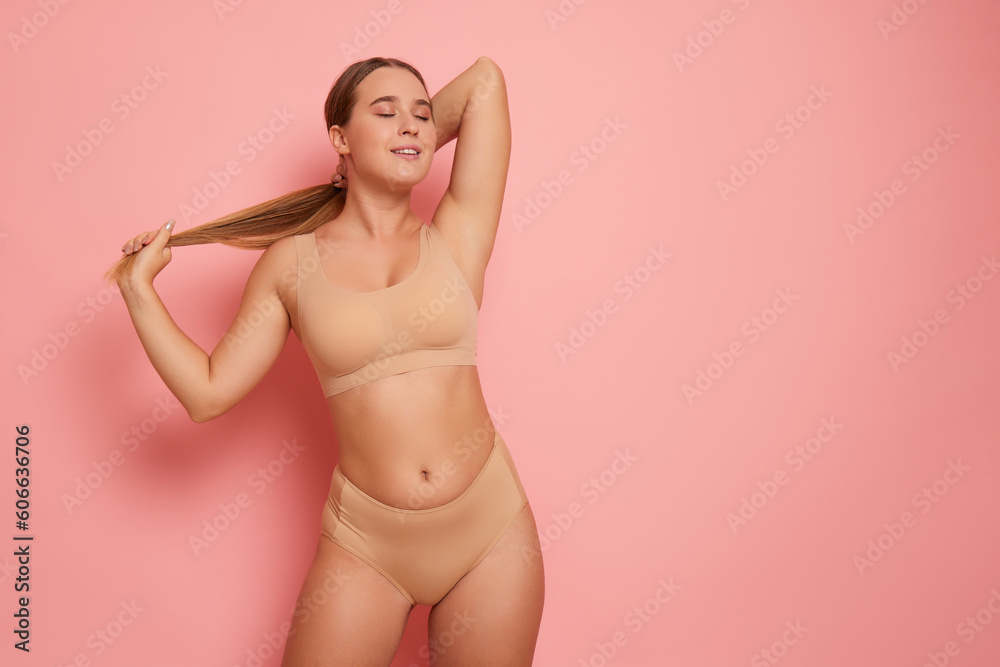 Pretty girl with long hair wearing beige colour bra and panties poses on pink background in sexy pose with hands on her hair, body comfort concept, copy space - obrazy, fototapety, plakaty 