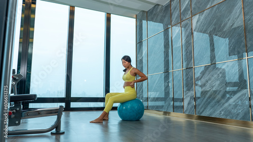Woman working out with fitness ball in gym, doing exercises for muscle press in white room, yoga meditates against modern skyscrapers. Young girl spending time fitness room on roof top of condominium