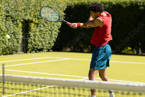 Biracial young man hitting ball with tennis racket while playing on grassy field at tennis court