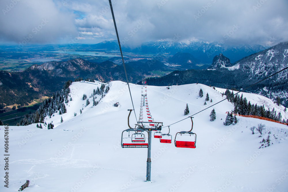 Empty red chairlift near the summit of Mount Breitenberg in the German Alps, surrounded by contrasting wintry landscape up high and green meadows in the valleys below