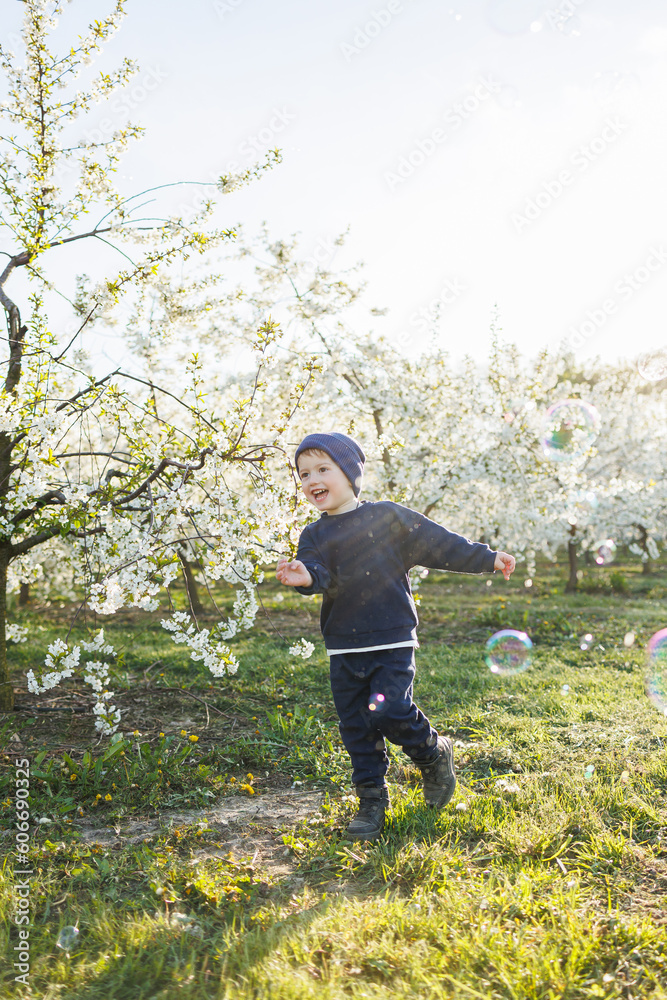 A little boy, 3 years old, in a sweater and a hat, runs through a blooming garden. Clothes for children aged 3 years. A happy eotian child is white among blossoming apple trees