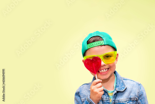 A seven year old cute boy with a beautiful smile and a green cap in light green glasses holds a big red heart lollipop in front of his eye covering it