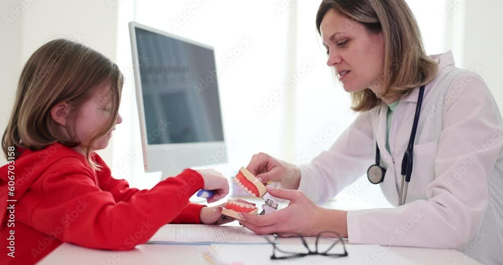 Girl client learns correct brushing teeth technique on jaw at white table in clinic premise. Orthodontist prompts girl explaining harm of improper cleaning slow motion Girl client learns correct brushing teeth technique on jaw at white table in clinic premise. Orthodontist prompts girl explaining harm of improper cleaning slow motion