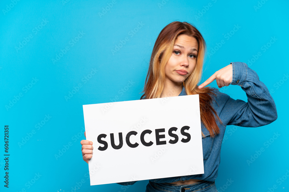 Teenager girl over isolated blue background holding a placard with text SUCCESS and pointing it Teenager girl over isolated blue background holding a placard with text SUCCESS and pointing it