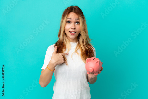 Teenager blonde girl holding a piggybank over isolated blue background with surprise facial expression