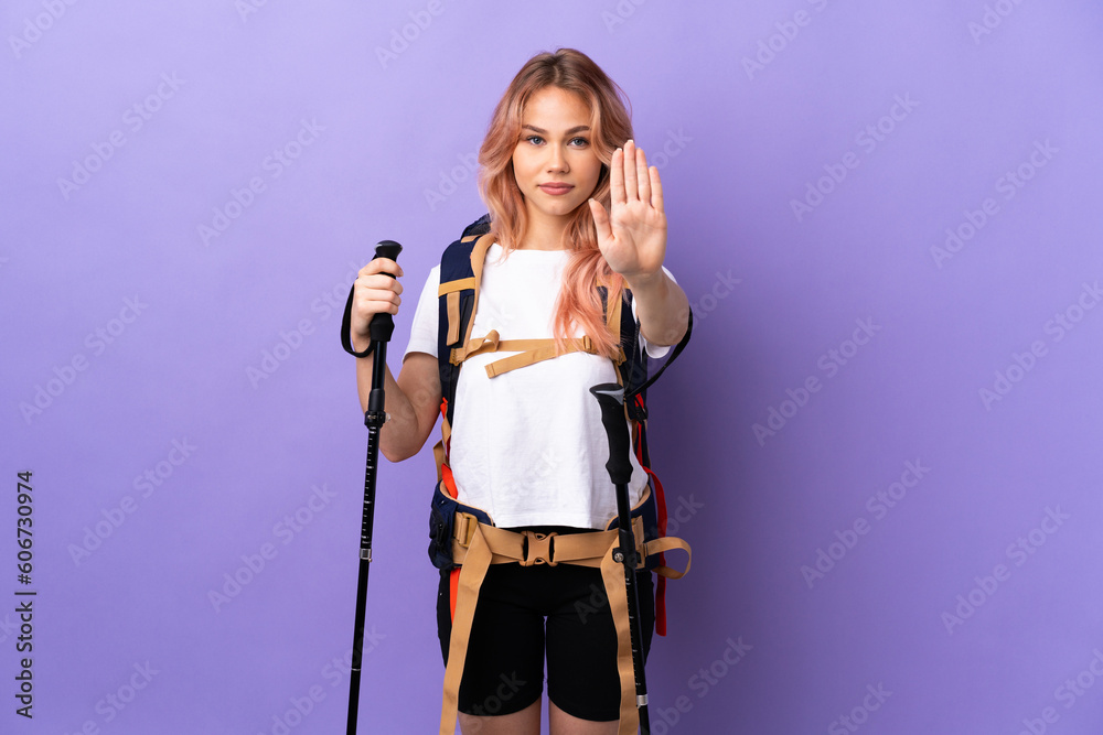 Teenager girl with backpack and trekking poles over isolated purple background making stop gesture Teenager girl with backpack and trekking poles over isolated purple background making stop gesture