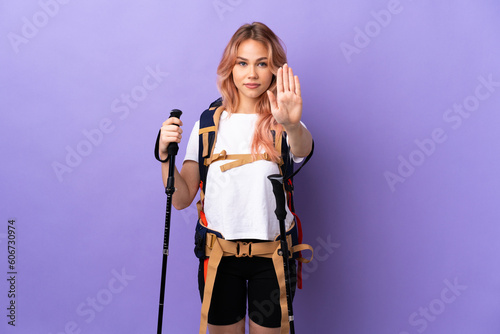 Teenager girl with backpack and trekking poles over isolated purple background making stop gesture