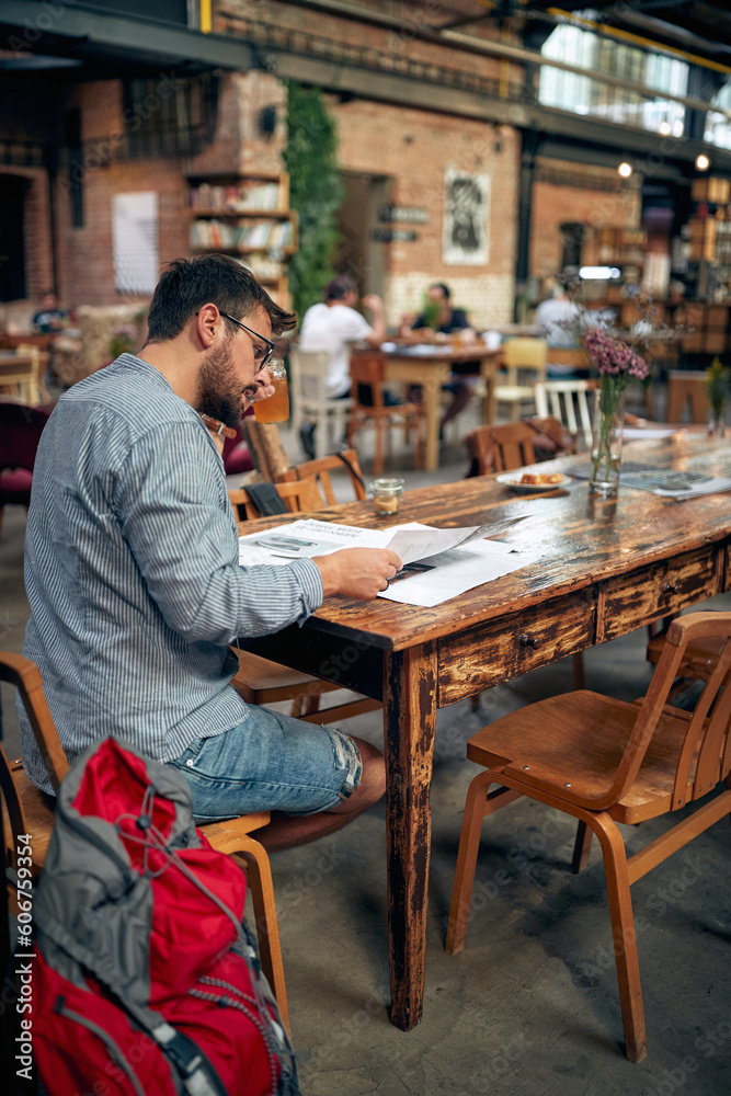 Young bearded man in casual wear with backpacker bag beside him, sitting alone at table reading newspaper. Young bearded man in casual wear with backpacker bag beside him, sitting alone at table reading newspaper.