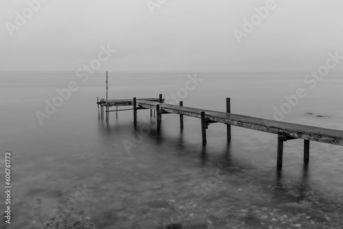 Grayscale shot of a wooden pier on a rocky shore against cloudy sky in Denmark