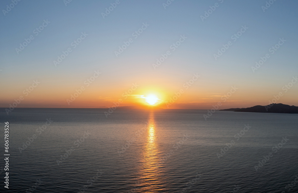 Sunset over Cyclades island Greece. Golden sun gets hide behind hill and colors gold sea water.