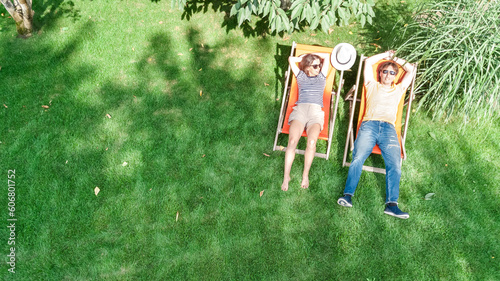Young couple relax in summer garden in sunbed deckchairs on grass, woman and men have drinks on picnic outdoors in green park on vacation, aerial top view from above
