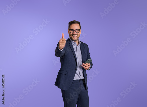Cheerful male entrepreneur gesturing thumbs up after reading good news over smart phone. Happy young businessman showing perfect and approval sign while standing isolated on blue background photo