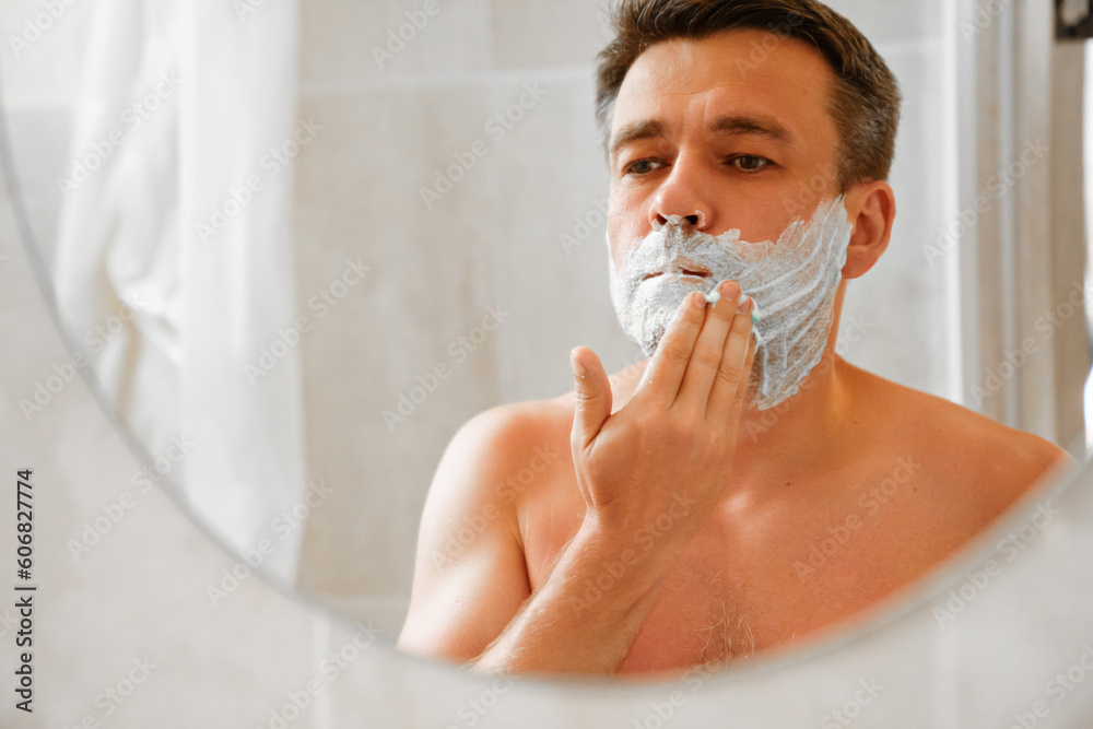a man applies shaving foam on his face and looks in a round mirror. The concept of men's cosmetics and shaving accessories. daily hygiene rituals.