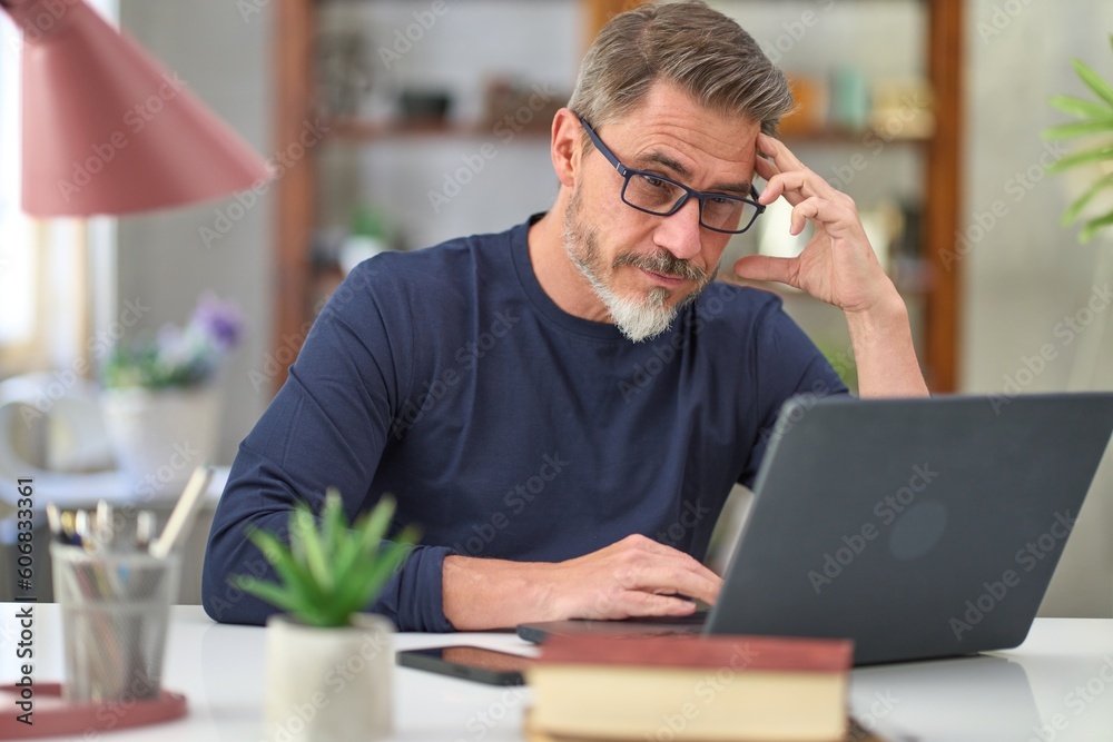 Casual mid adult man with laptop computer at desk in home office, banking online, remote working. Portrait of older gray haired bearded guy thinking. Businessman managing business on internet. Casual mid adult man with laptop computer at desk in home office, banking online, remote working. Portrait of older gray haired bearded guy thinking. Businessman managing business on internet.