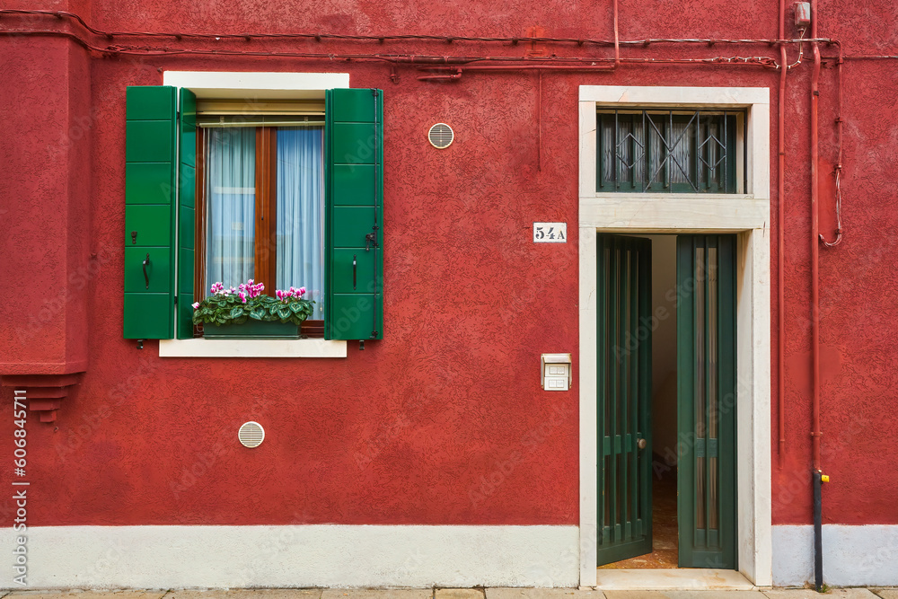 Door and window with flower on the red facade of the house. Colorful architecture in Burano island, Venice.