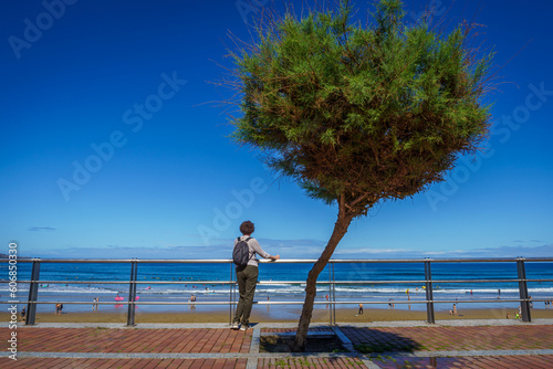 Einsame Frau mit Rucksack steht neben einer Tamariske auf der Promenade von Las Palmas de Gran Canaria und beobachtet Menschen am Strand
