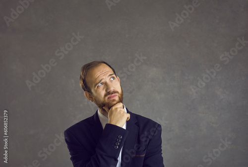 Fototapeta Naklejka Na Ścianę i Meble -  Portrait of puzzled and confused man with pensive facial expression against gray background. Bearded man touching his chin pondering what choice to make. Decision making concept. Banner.