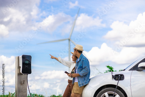 Concept of progressive happy young couple enjoying their time at windmill farm with electric vehicle. EV car driven by clean renewable energy from wind turbine generator for charging station. photo