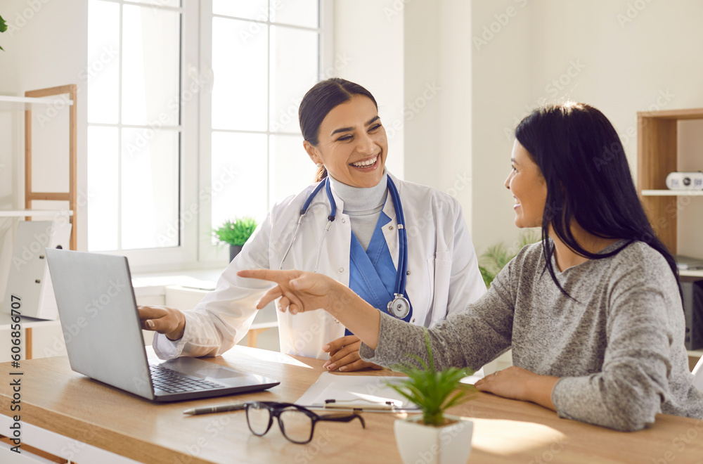 Female young woman patient visiting a smiling friendly doctor at the medical office in hospital, looking at the laptop monitor screen and having consultation during medical examination in clinic. Female young woman patient visiting a smiling friendly doctor at the medical office in hospital, looking at the laptop monitor screen and having consultation during medical examination in clinic.
