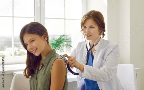 Female pediatrician listens to breathing of preteen girl, applying stethoscope to her back. Young friendly woman listens to presence of wheezing in lungs of patient's child during medical examination. photo