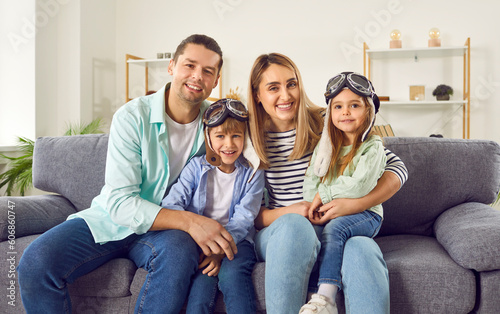 Portrait of happy smiling married couple sitting on sofa in the living room at home with their two kids boy and girl in pilot's glasses and looking at camera cheerfully. Family and people concept.