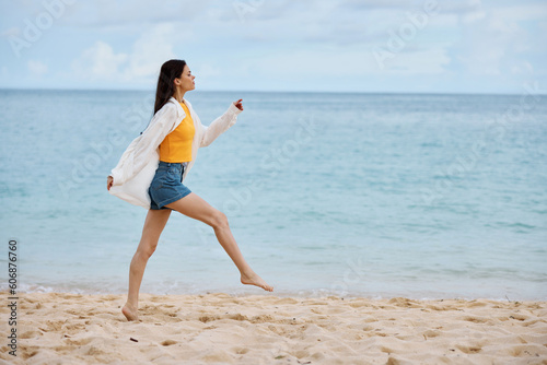 Fototapeta Naklejka Na Ścianę i Meble -  Sports woman runs along the beach in summer clothes on the sand in a yellow tank top and denim shorts white shirt flying hair ocean view, beach vacation and travel